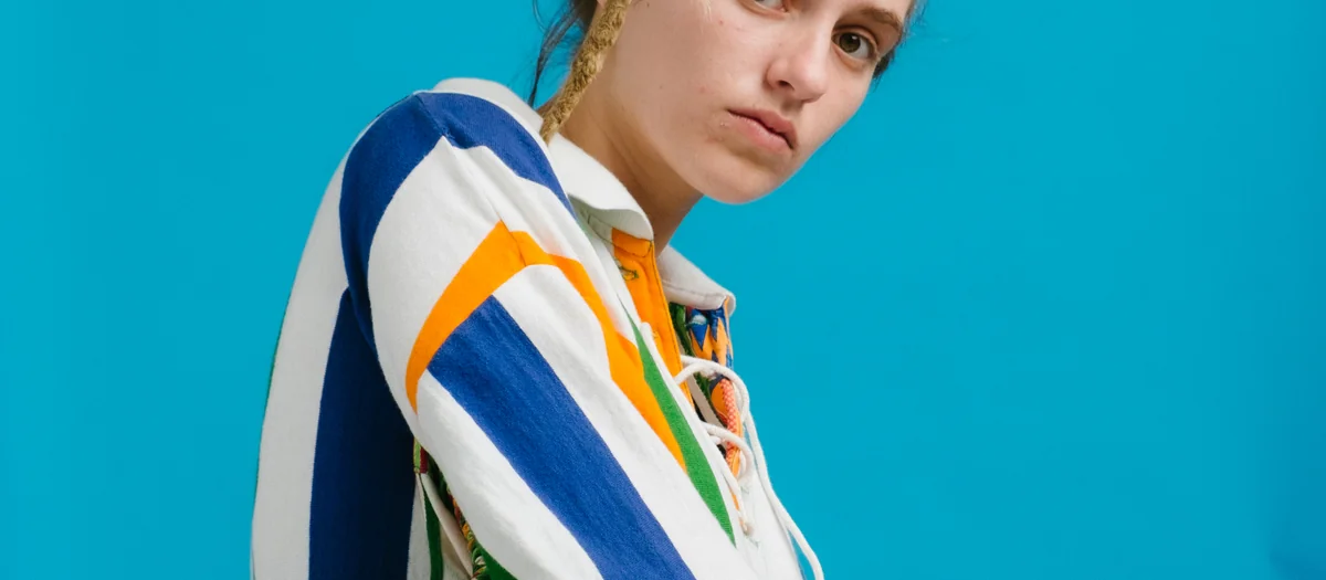 Model with blonde dreadlocks in a striped rugby shirt holding a striped bag against a blue backdrop