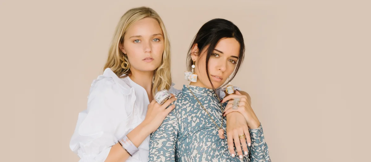 Two models posed together, one in a white ruffled blouse and one in a blue and white print top, on a beige backdrop