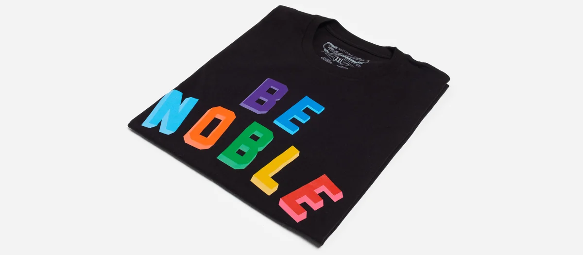 Folded black graphic tee with multicolor BE NOBLE letters photographed as a product flat on a light gray background
